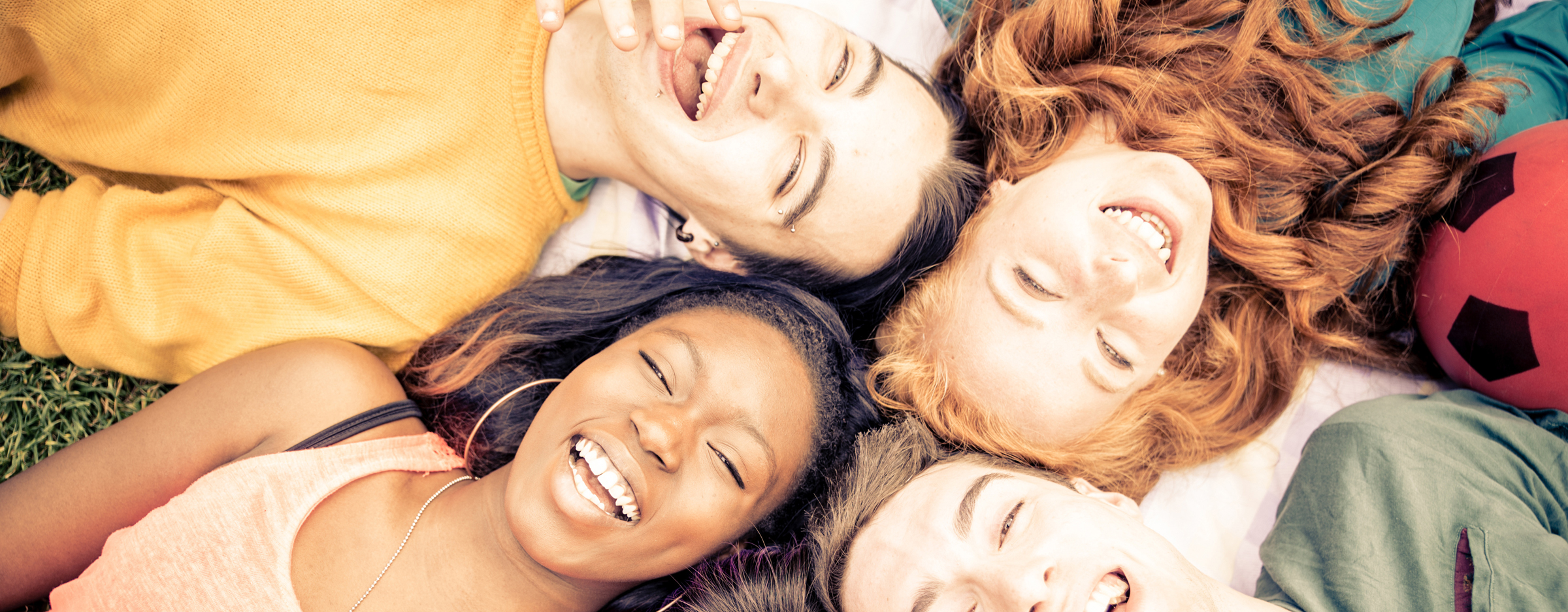 Fünf junge Menschen liegen mit ihren Köpfen im Kreis auf einer Wiese und lachen.