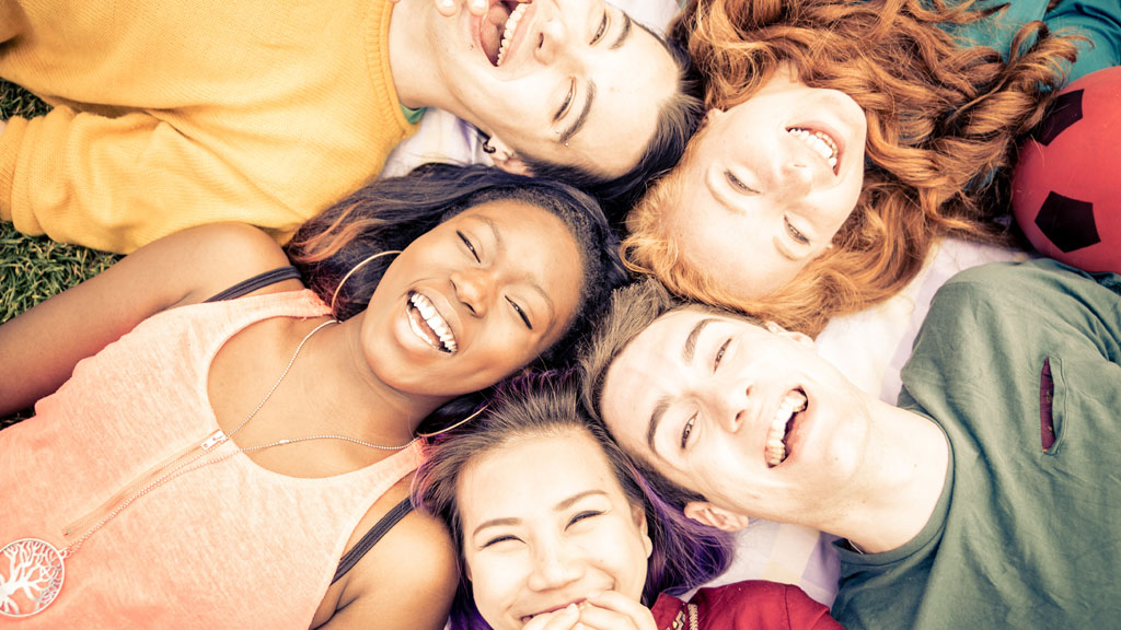 Fünf junge Menschen liegen mit ihren Köpfen im Kreis auf einer Wiese und lachen.