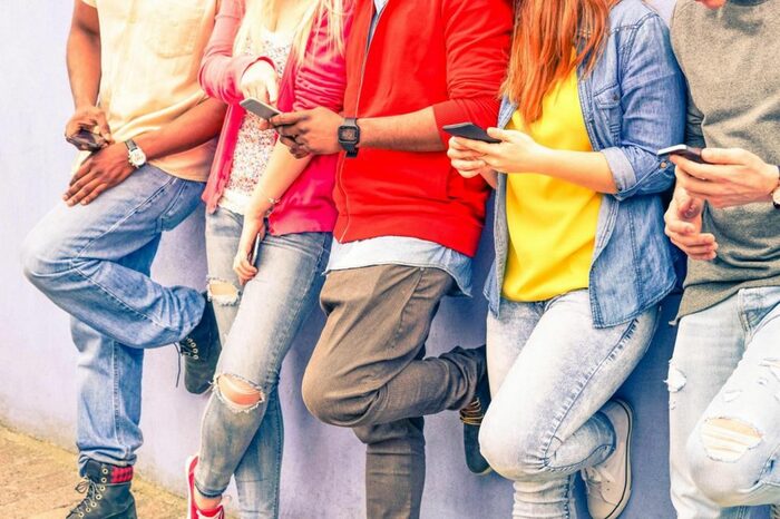 Fünf Jugendliche in Jeans stehen vor Wand mit Smartphones in der Hand